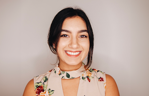 Headshot of Avani. She is wearing a floral top and is standing in front of a light grey background.