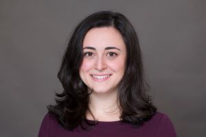 Headshot of Jackie. She is wearing a burgundy top and is standing in front of a dark grey background.