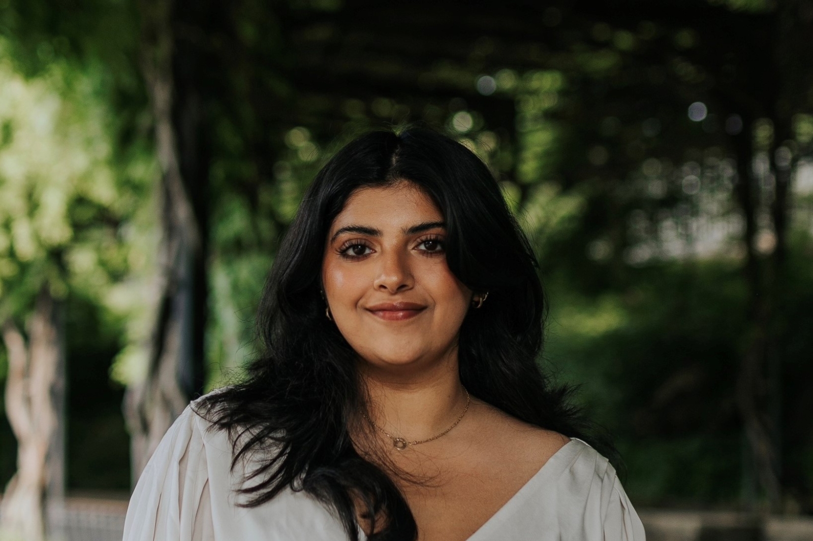 Headshot of Sara. She is wearing a white top and is standing in front of a garden background with various vegetation.