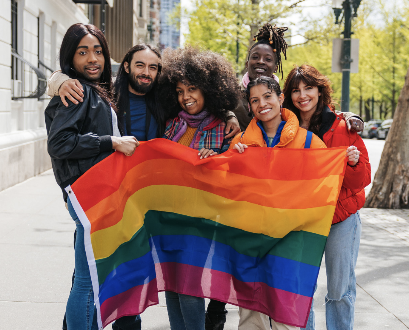 6 people are standing on the sidewalk and are collectively holding one pride flag. They all are smiling.