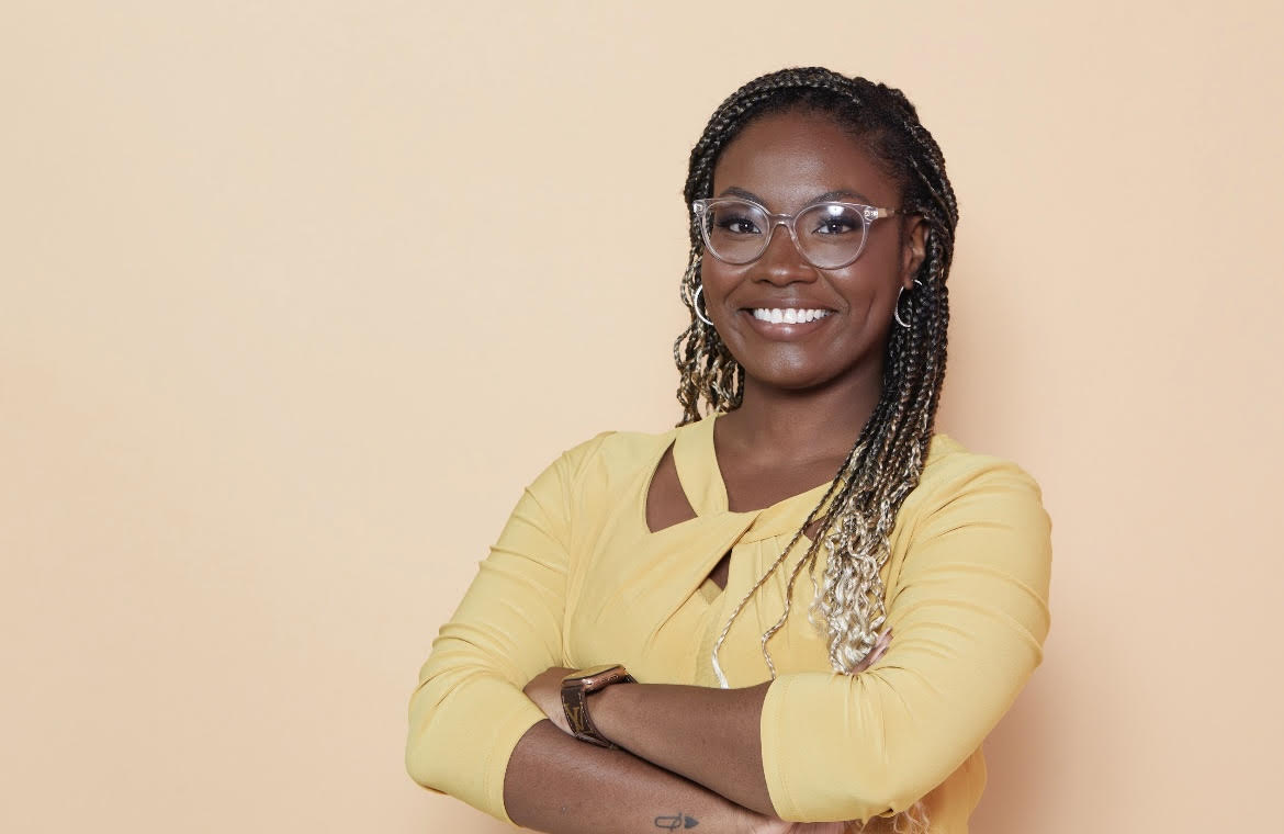 Headshot of Faith. She is wearing a yellow top and she is standing in front of a peach/tan background.