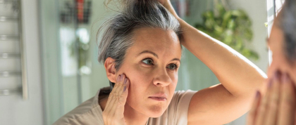 woman looks at greying hair and skin in mirror